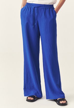 Kobieta ubrana w królewskie niebieskie spodnie o szerokich nogawkach z elastycznym pasem i troczkiem, w połączeniu z czarnymi sandałami z odkrytymi palcami.