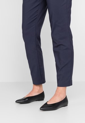 Osoba nosząca granatowe zwężane spodnie i czarne płaskie buty z lekko fakturowanym wzorem, stojąca na jednolitej podłodze.