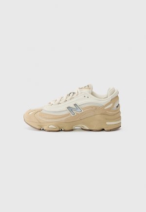Beżowo-białe tenisówki New Balance 1000 z szarym logo „N”, sznurowane z przodu i wyściełaną podeszwą na jednolitym jasnym tle.