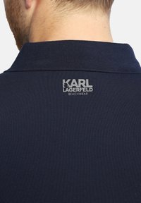 Marineblaues Polo-Shirt aus strukturiertem Stoff, mit Kragen und einem silbernen Logo "KARL LAGERFELD BEACHWEAR" auf der Rückseite.
