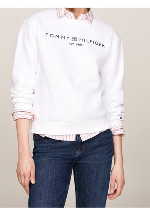 Person trägt weißen Tommy Hilfiger Sweatshirt über rosa gestreiftem Hemd und blauen Jeans, steht vor schlichtem Hintergrund.