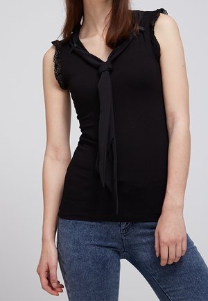 Frau trägt ein ärmelloses schwarzes Oberteil mit Spitzenbesatz und einem gebundenen Schal-Detail, kombiniert mit blauen Jeans, steht vor einem schlichten Hintergrund.