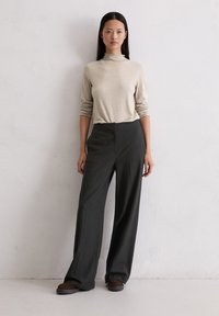 Beżowy sweter z golfem w połączeniu z ciemnoszarymi spodniami o szerokich nogawkach w prążki. Model stoi na jasnym tle. Brązowe buty dopełniają stylizację.