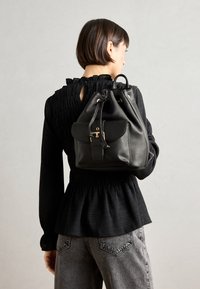 Sac en cuir noir en forme de seau avec fermeture à cordon et poche avant à rabat, avec des accents en métal doré. Porté sur le dos.