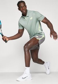 Light green polo shirt with a yellow logo, paired with grey athletic shorts. White sneakers with black accents. Active pose with a tennis racket.