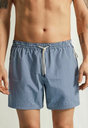 Homme portant un short de bain rayé vertical bleu et blanc avec un cordon blanc et une poche zippée sur le côté droit.