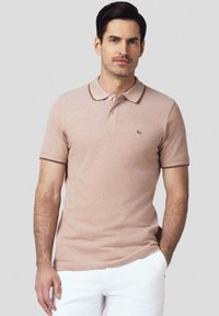 Koszulka polo w jasnoróżowym kolorze z prążkowanym kołnierzykiem i brązowym wykończeniem. Posiada logo na piersi oraz krótkie rękawy, wykonana z miękkiego, teksturowanego materiału.