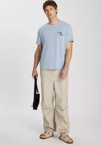 Jasnoniebieska koszulka z okrągłym dekoltem z małym nadrukiem na lewej piersi, w połączeniu z luźnymi beżowymi spodniami i brązowymi sandałami.