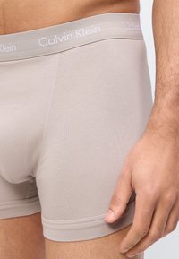 Μπεζ ανδρικά εσώρουχα boxer Calvin Klein σε άνδρα με το ένα χέρι χαλαρά δίπλα του, δείχνοντας την υφή του υφάσματος και τη ζώνη μέσης με το λογότυπο.