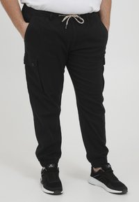 Schwarze Cargo-Hosen mit elastischem Bund, Kordelzug und seitlichen Taschen, ausgestattet mit elastischen Bündchen und einer glatten, matten Stofftextur.