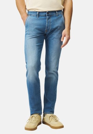 Uomo che indossa sneakers beige chiaro e jeans slim-fit blu medio con camicia bianca parzialmente visibile, in piedi davanti a uno sfondo grigio.