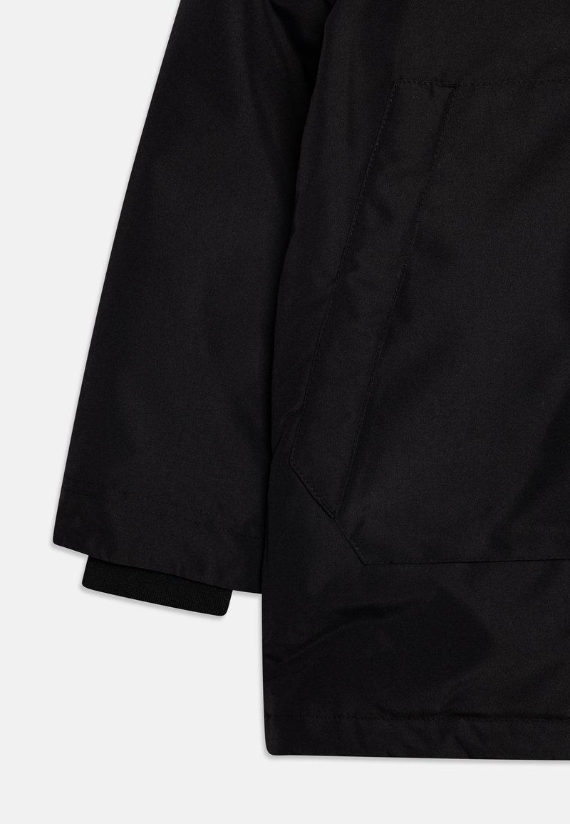 Veste extérieure noire en tissu texturé, avec des poignets élastiques et une poche latérale aux bords angulaires.