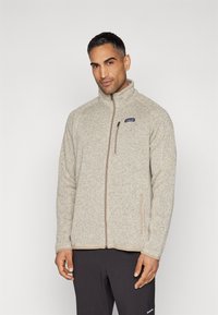 Giacca in pile beige chiaro con colletto alto, chiusura con zip e due tasche laterali. Presenta un piccolo logo sul lato sinistro del petto.