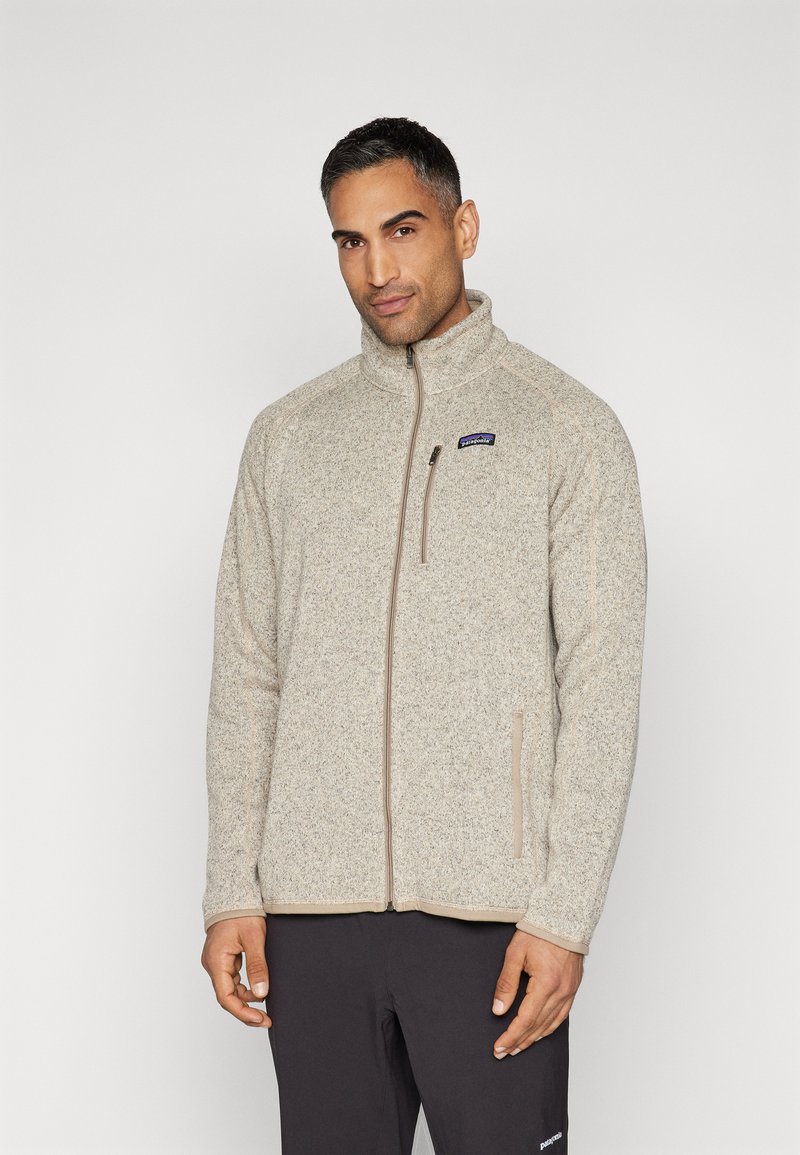 Giacca in pile beige chiaro con colletto alto, chiusura con zip e due tasche laterali. Presenta un piccolo logo sul lato sinistro del petto.