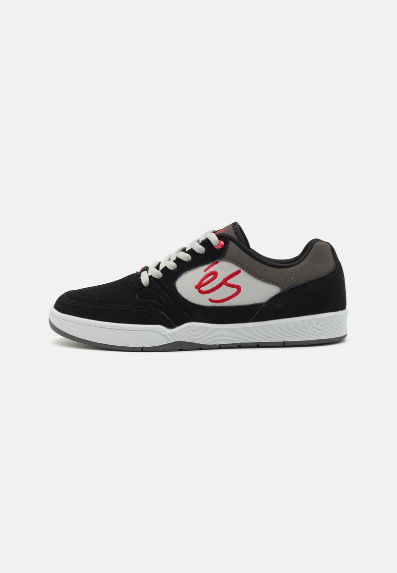 Des chaussures de skate en suede noir avec des accents blancs et gris, arborant un logo rouge, des lacets plats et une semelle en caoutchouc blanc.