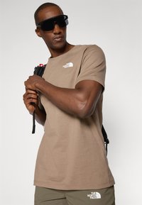 Tricou din bumbac maro cu guler rotund, având un logo alb pe piept și mâneci scurte. Ochelari de soare negri purtați, ținându-se de bretele.