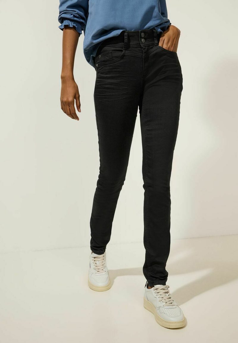 Jeans ajustados negros de mezclilla, con una cintura alta, dos bolsillos laterales y dos bolsillos traseros, combinados con zapatillas blancas.