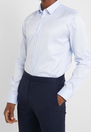 Homme portant une chemise bleu clair boutonnée rentrée dans un pantalon formel bleu marine foncé avec une main dans la poche.