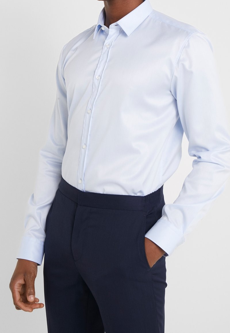 Homme portant une chemise bleu clair boutonnée rentrée dans un pantalon formel bleu marine foncé avec une main dans la poche.