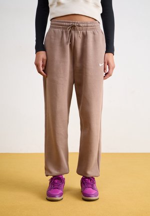 Personne portant un pantalon de survêtement Nike marron avec cordon de serrage, un haut court crème et noir à manches longues, et des baskets rose vif debout sur un sol jaune.