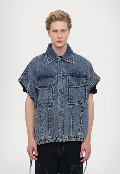 Denimová overshirt s krátkými rukávy, vybavená dvěma velkými předními kapsami, zapínáním na knoflíky a nařaseným lemem. Nositelná přes černý top.