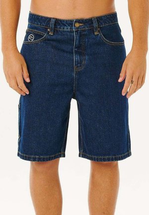 Homme portant un short en denim bleu foncé avec des poches avant et un logo sur la poche droite, debout devant un fond uni.