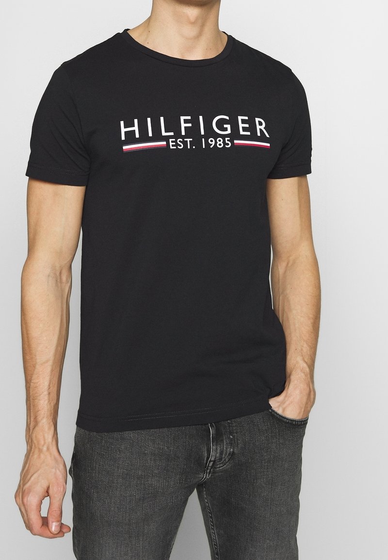 Black cotton T-shirt featuring a crew neck, short sleeves, and front white lettering with red and blue stripes. Pairs with dark jeans.