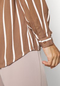 Blouse à rayures marron avec des rayures verticales blanches. Tissu doux, détails de poignets boutonnés et coupe décontractée. Portée avec un pantalon beige clair.