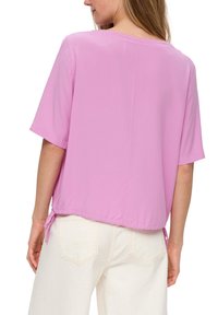 Blouse rose à manches courtes avec une coupe ample et un cordon de serrage à l'ourlet. Texture lisse et encolure ronde. Portée avec un pantalon de couleur claire.