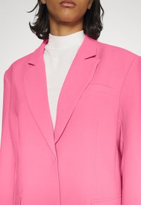 Blazer fúcsia ajustado com lapelas afiadas e um único bolso no peito, sobreposto a um top branco de gola alta. Tecido liso e texturizado.
