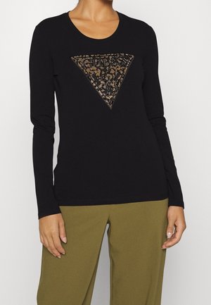 Osoba ubrana w czarną koszulę z długim rękawem z logo Guess w panterkę oraz oliwkowe spodnie, stojąca z luźno opuszczonymi ramionami.