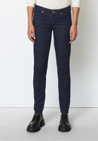 Dark blue denim skinny jeans with a mid-rise waist, two front pockets, and tan stitching. Paired with black lace-up platform shoes.