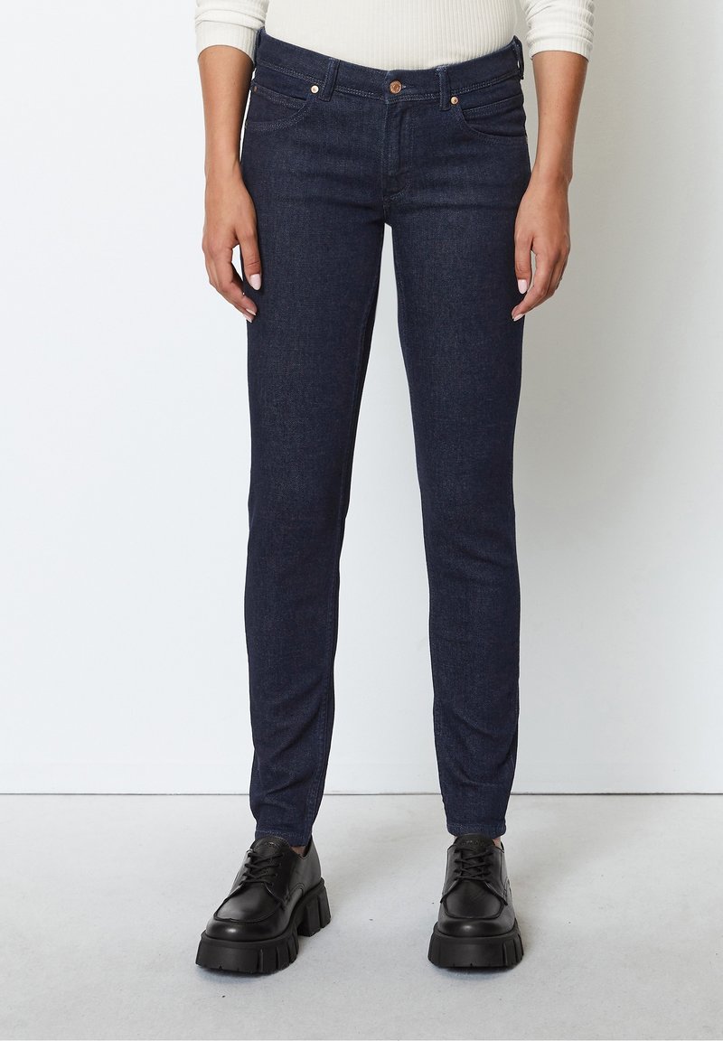 Dark blue denim skinny jeans with a mid-rise waist, two front pockets, and tan stitching. Paired with black lace-up platform shoes.