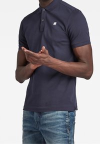 Marinblå pikétröja med krage, två knappar och en liten logotyp; tillverkad av mjukt tyg, smal passform, kombinerad med blå jeans.