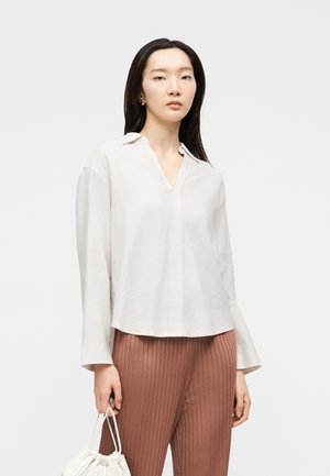 Femme aux longs cheveux noirs portant un chemisier blanc à col et un pantalon côtelé marron, tenant un petit sac à main blanc sur un fond uni.