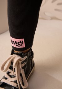 Baskets montantes noires avec coutures blanches, lacets crème et une étiquette en tissu rose portant l'inscription "BLUEY" à la cheville de leggings noirs côtelés.