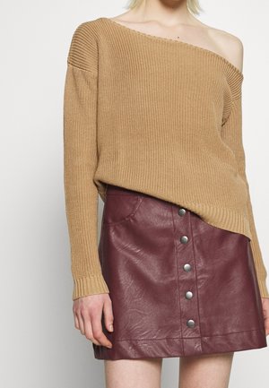 Femme portant un pull en maille beige à épaules dénudées et une mini-jupe en cuir bordeaux avec boutons sur le devant, montrant le haut du torse et les hanches.