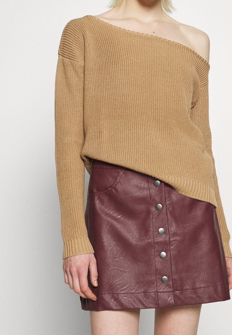 Femme portant un pull en maille beige à épaules dénudées et une mini-jupe en cuir bordeaux avec boutons sur le devant, montrant le haut du torse et les hanches.