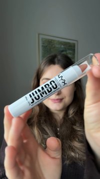 Persoon die de NYX Jumbo multifunctionele gezichtsmake-upstick met een doorzichtige dop vasthoudt, met de focus op de producttekst en de punt tegen een vervaagde achtergrond.