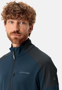 Giacca zip-up navy in tessuto testurizzato, con pannelli neri sulle spalle e logo riflettente. Collo alto e dettagli di chiusura a zip frontale.