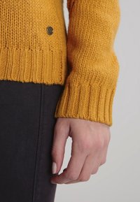 Senfgelber Strickpullover mit geripptem Bündchen; verfügt über einen kleinen metallischen Akzent an der Seite und wird mit einer dunkel gefärbten Hose kombiniert.