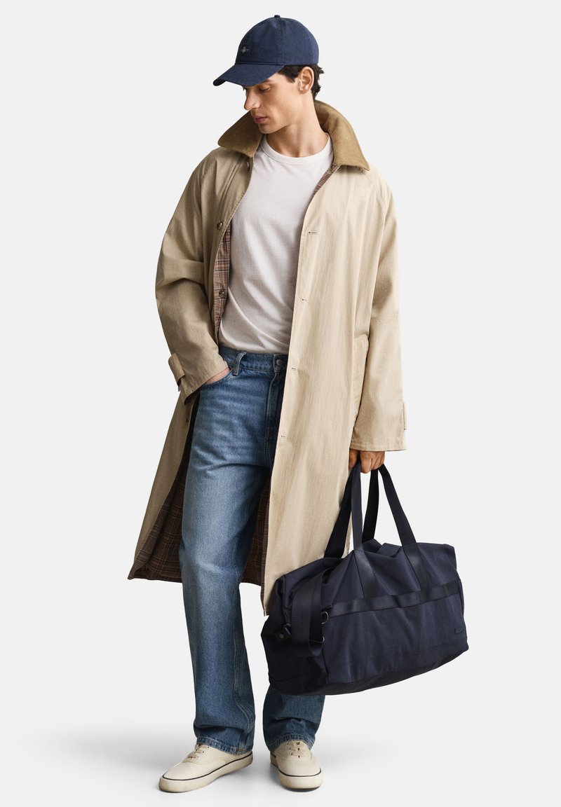 Άνδρας που φοράει μακρύ μπεζ παλτό, λευκό μπλουζάκι, μπλε τζιν, μπλε σκούφο, κρατώντας με το ένα χέρι μια μεγάλη μπλε τσάντα duffel.