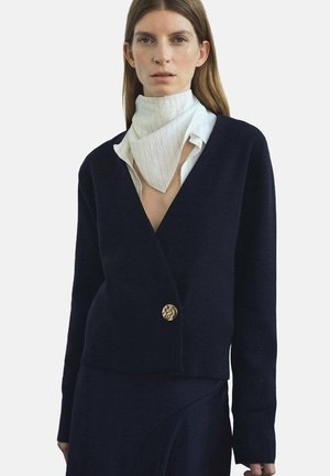 Marineblauer Strickcardigan mit tiefem V-Ausschnitt, langen Ärmeln und einem goldenen Knopfdetail an der Taille. Über einem weißen Hemd mit feinen Streifen gestylt.