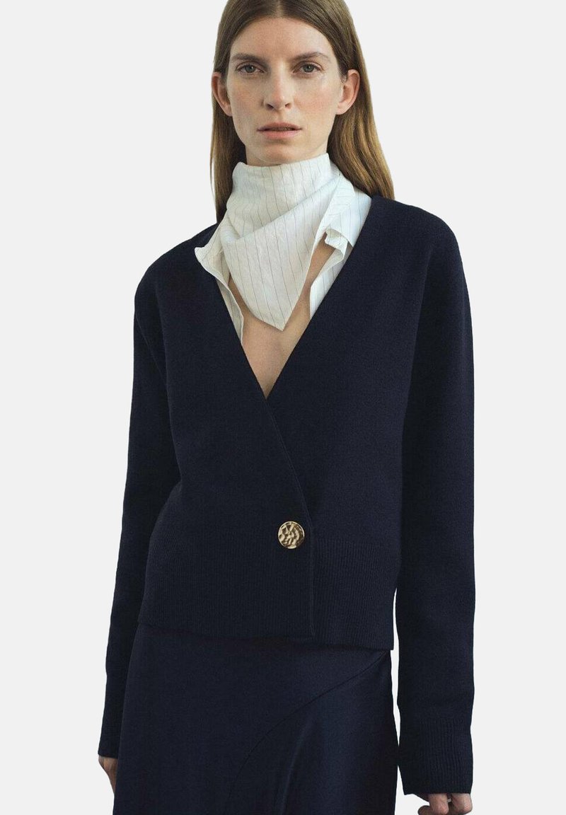 Námornícky pletený kardigán s hlbokým V-výstrihom, dlhými rukávmi a zlatým gombíkom v oblasti pása. Uložený na bielom košeli s pásikmi.