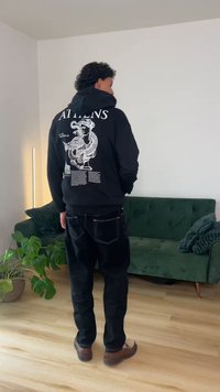 Άνδρας που βρίσκεται σε δωμάτιο, φοράει μαύρο hoodie με λευκό σχέδιο και κείμενο Αθηνά, μαύρο παντελόνι, καφέ παπούτσια, κοντά σε πράσινο καναπέ και φυτά.