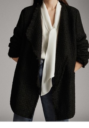 Donna che indossa un cardigan nero testurizzato aperto sul davanti sopra una camicetta bianca con collo annodato, abbinata a jeans blu scuro, con le mani nelle tasche.