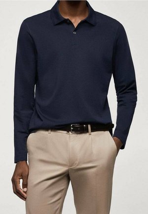 Mand iført marineblå langærmet poloshirt stukket ned i beige bukser med sort bælte, hånd i lommen, mod en ensfarvet baggrund.