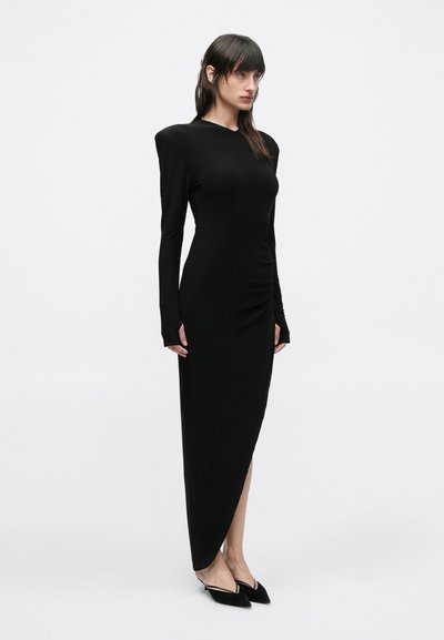 Μαύρο φόρεμα με μακριά μανίκια και ασύμμετρο τελείωμα. Λείο, ελαστικό ύφασμα, με δομημένους ώμους και διακριτική ραφή στη μέση. Μαύρα μυτερά παπούτσια.