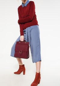 Femme tenant un sac à main bordeaux, portant un pull rouge par-dessus une chemise bleu à col, des culottes bleues et des bottines à talons couleur rouille.