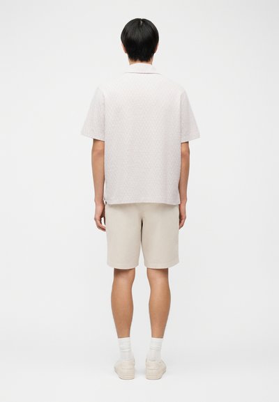 Άνδρας που στέκεται γυρισμένος προς τα πίσω, φοράει ανοιχτό μπεζ κοντομάνικο πουκάμισο, μπεζ σορτς, άσπρες κάλτσες και μπεζ αθλητικά παπούτσια, μπροστά σε απλό φόντο.